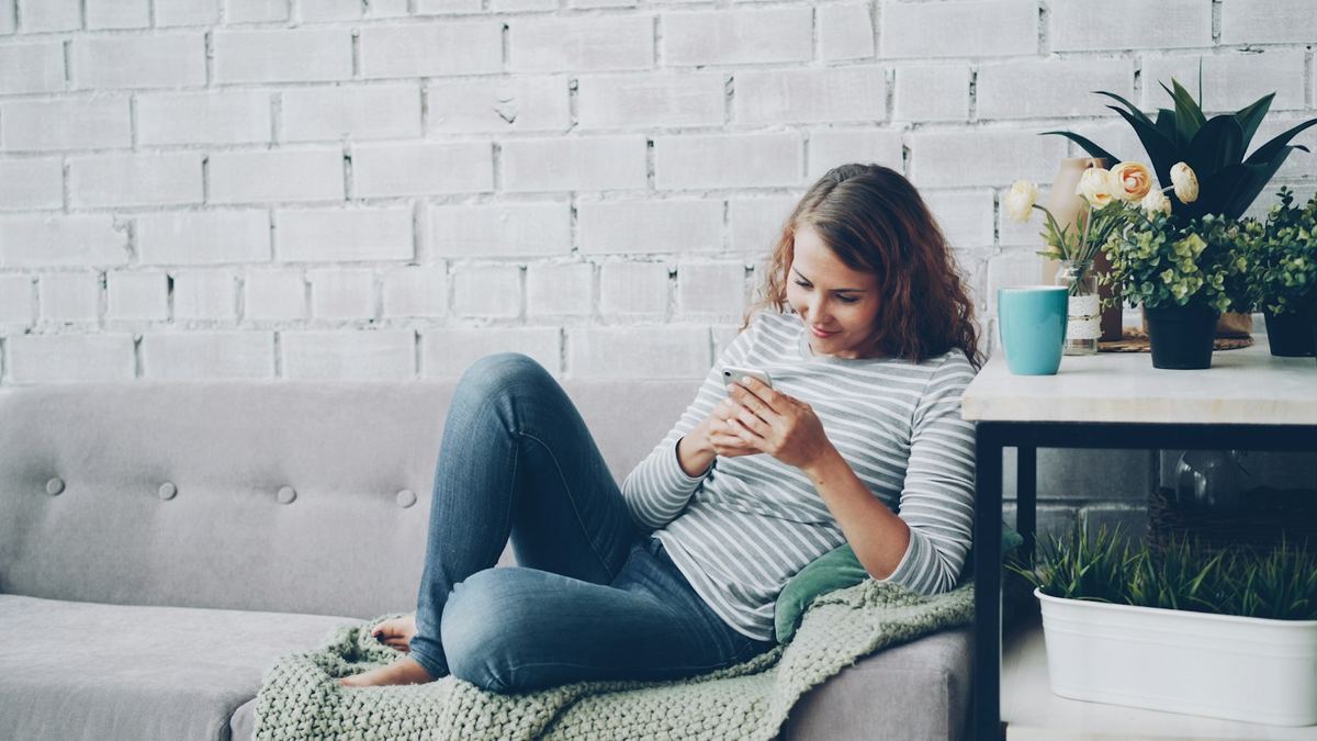 Jovem sentada no sofá do apartamento usando o celular